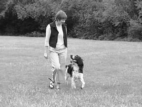 A woman walks in a grassy field with a dog walking at her side