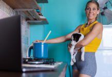 Woman, with a small dog in her arms cooking at stove while watching recipe on laptop.