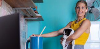 Woman, with a small dog in her arms cooking at stove while watching recipe on laptop.