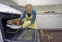 A older woman pulls a turkey out of the oven while a small dog looks on in anticipation.