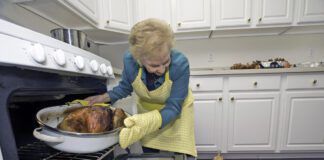 A older woman pulls a turkey out of the oven while a small dog looks on in anticipation.