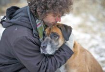 Young Caucasian man wearing a winter coat and a scarf hugging his pet dog lovingly to comfort him as they are out for a walk through a snowy forest during winter. The dog is a mixed breed and brown and black in color with a sad expression on his face.