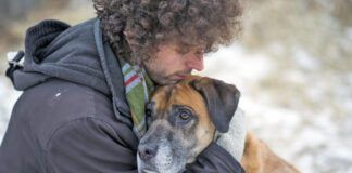 Young Caucasian man wearing a winter coat and a scarf hugging his pet dog lovingly to comfort him as they are out for a walk through a snowy forest during winter. The dog is a mixed breed and brown and black in color with a sad expression on his face.